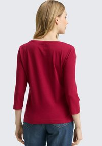 Rotes, langärmliges Oberteil mit rundem Halsausschnitt und glatter Textur, kombiniert mit blauen Jeans, von hinten betrachtet.