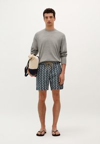 Chemise grise à manches longues, shorts de bain bleus avec un motif de vagues, et tongs noires. Tient un sac beige et marine.