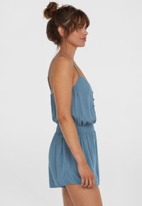 Blue sleeveless playsuit with a smocked waist, textured fabric, button details down the front, and a relaxed fit.