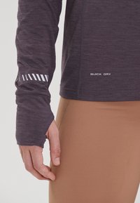 Purple long-sleeve top with a textured finish and reflective white stripes on the sleeve; paired with light brown fitted leggings.
