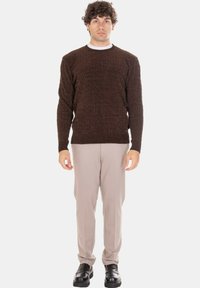 Giovane uomo in piedi che indossa un maglione marrone lavorato a maglia, pantaloni beige e scarpe di pelle nere su uno sfondo chiaro e uniforme.