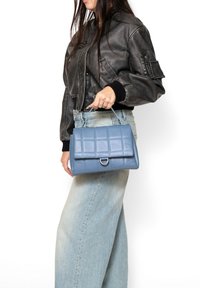 Borsa in pelle blu con motivo trapuntato, forma rettangolare e accessori metallici argentati. Tenuta da una persona con una giacca bomber nera.
