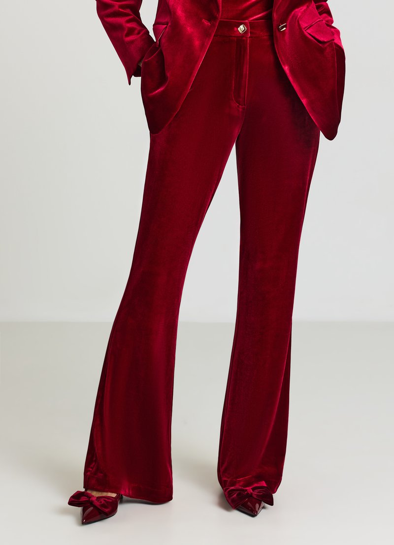 Pantalon évasé en velours rouge avec une texture lisse. Il présente une taille haute, des poches latérales et des accents de noeuds aux ourlets. À associer avec un blazer assorti.
