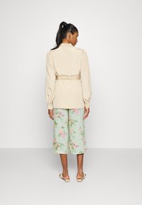Blusa de cetim bege com mangas bufantes, cintura ajustada, combinada com calças largas de estampado floral em verde pastel, e sandálias planas.