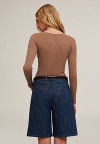 Geribbelde bruine lange mouw top gecombineerd met knielange blauwe denim shorts. De shorts hebben een klassieke vijf-pocketstijl en een donkere riem.