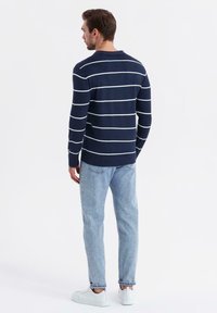 Muž s hnědými vlasy, oblečený v námořnickém svetr s bílými pruhy, světle modrých džínách a bílých teniskách, stojící zády k bílému pozadí.