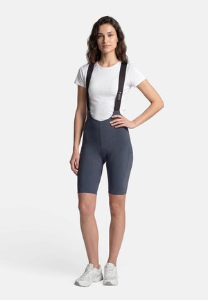 Femme portant un t-shirt blanc, un cuissard de cyclisme gris foncé avec des bretelles noires et des baskets blanches, debout avec une main sur la hanche.