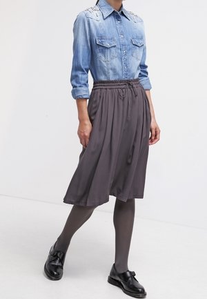 Woman wearing blue denim shirt with pearl details, gray midi skirt, dark tights, and black buckle shoes walking against white background.