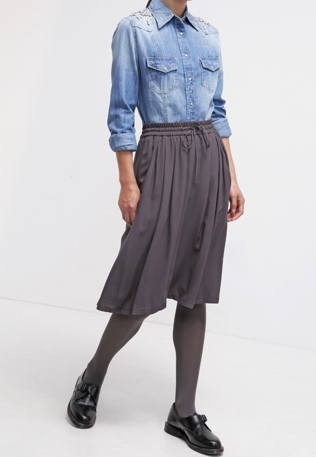 Woman wearing blue denim shirt with pearl details, gray midi skirt, dark tights, and black buckle shoes walking against white background.
