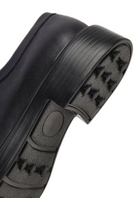 Scarpa nera con suola in gomma texturizzata caratterizzata da tacchetti di presa rialzati e una parte superiore liscia, progettata per stabilità e aderenza.