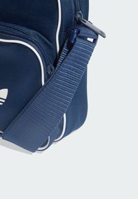 Prekrižna torbica tamnoplave tkanine s bijelim obrubom, teksturiranim remenom i pretincem na zatvarač, koja prikazuje minimalistički dizajn i praktične značajke.