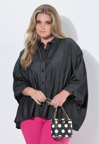 Frau mit langen blonden Haaren trägt eine lockere, dunkle Bluse mit Knöpfen und leuchtend pinke Hose. Sie hält eine kleine Handtasche mit schwarz-weißen Punkten und einer goldenen Kette.