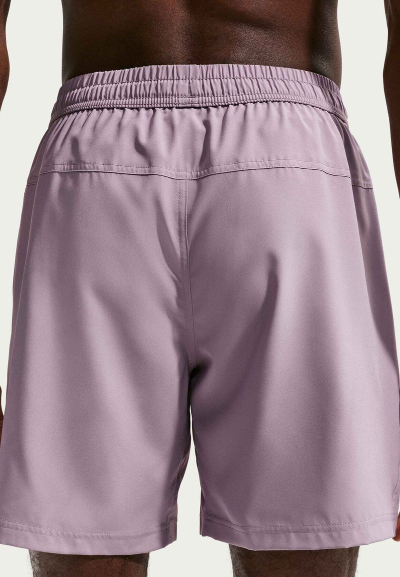 Homme de la taille vers le bas portant un short violet clair à taille élastique, debout devant un fond uni clair.