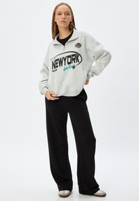 Grå sweatshirt med hög krage, prydd med svarta och teal grafiker som står "NEW YORK". Matchad med svarta vida byxor och beige skor.