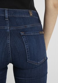 Jeans de mezclilla de color azul oscuro con un diseño ajustado, que cuentan con dos bolsillos traseros, costuras en contraste y una pequeña etiqueta de marca en la cintura.