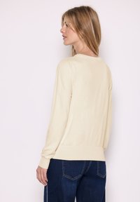 Maglione beige a maniche lunghe con scollatura rotonda e polsini a coste, abbinato a jeans a gamba larga di colore blu scuro, con una texture liscia.