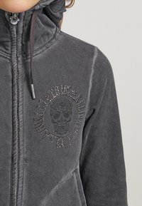 Haut à zip gris avec un col montant, présentant un graphique de skull avec des accents de strass sur le devant. Fabriqué en tissu doux avec des coutures visibles.