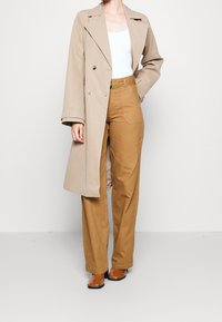 Žena v béžovém trenčkotě, bílé tílku, hnědých vysokých širokých kalhotách a hnědých kožených botách, pózující s jednou rukou v kapse.