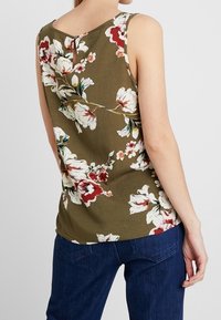 Débardeur floral en tissu vert olive à motifs, avec de grandes fleurs blanches et rouges. Le dos présente un détail en forme de trou de serrure.