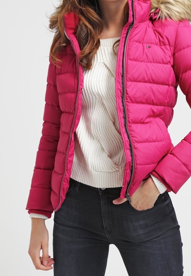 Kobieta ubrana w jasnoróżową pikowaną kurtkę z kapturem obszytym futerkiem, białym prążkowanym swetrem i ciemne dżinsy, z ręką w kieszeni kurtki.