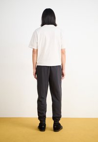 Ljusgrå kortärmad skjorta i kombination med mörkgrå mjukisbyxor och svarta skor, sedd bakifrån på en gul yta.