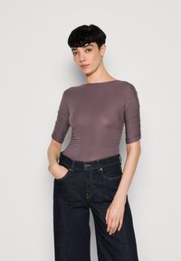 Mauve topný bodysuit s třičtvrtinovými rukávy a řasením, spojený s vysokým pasem tmavě modrými širokými džínami.