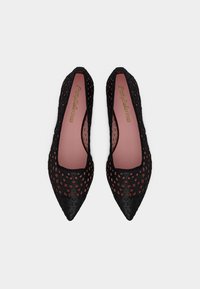 Ζευγάρι μαύρα φλατ παπούτσια με μυτερή μύτη και γεωμετρικές εγκοπές, με ροζ εσώσολες, τοποθετημένα δίπλα δίπλα σε λευκό φόντο.