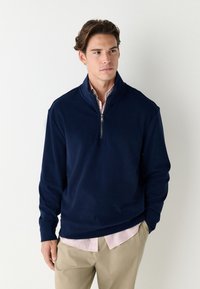 Námořnický pulovr s čtvrtým zipem vyrobený z měkké látky, s dlouhými rukávy a žebrovaným lemem. Nošený přes světle růžovou a bílou pruhovanou košili.