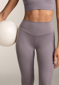 Högmidjade lavendelfärgade leggings med en slät textur, tillsammans med en croppad topp, hållna mot en vit texturerad boll. Enkel design, inga mönster.