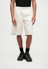 Shorts de couleur crème avec une taille élastique et un cordon de serrage, dotés d'ouvertures latérales et d'une texture lisse et douce. Portés avec des chaussettes noires et des baskets.