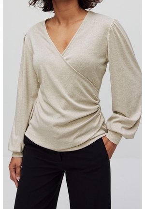 Kvinde iført en beige langærmet slå-om-top med let glimmer og sorte bukser, den ene hånd i lommen, foran en ensfarvet baggrund.