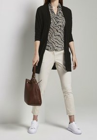 Schwarzer, langer Cardigan über einer schwarz-weißen, wellenförmig gemusterten Bluse, kombiniert mit hellbeigen Hosen und weißen Sneakers. Braune Leder-Tote.