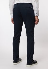 Marineblaue Slim-Fit-Hose mit glatter Textur, zwei Gesäßtaschen und einem Reißverschlussdetail. Kombiniert mit schwarzen Sneakers.