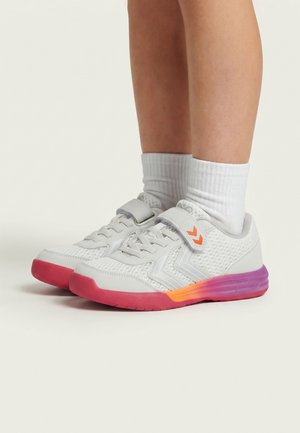 Dziecko noszące białe trampki z różowo-fioletowymi podeszwami i pomarańczowym logo, w połączeniu z białymi skarpetkami na gładkim tle.