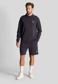 Marineblauwe rits-polo met een groen "LS" logo, gecombineerd met bijpassende shorts. Heeft een comfortabele pasvorm, kraag en ribgebreide manchetten. Beige sneakers en witte sokken.