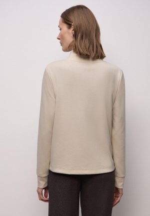 Felpa beige a collo alto, tessuto morbido, maniche lunghe, design aderente. Indossata con pantaloni marrone scuro a motivo, con le mani appoggiate di lato.