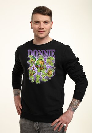 Mand iført sort sweatshirt med farverig Teenage Mutant Ninja Turtles-figur Donatello og teksten "DONNIE" foran.