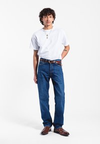 Camiseta blanca, jeans de mezclilla azules, cinturón de cuero marrón y zapatos marrones. El modelo está de pie en un entorno neutro, con cabello rizado y tatuajes visibles.