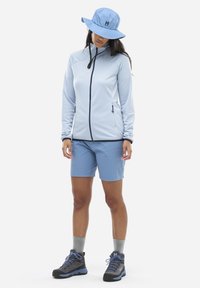 Hellblaues Reißverschlussjackett mit einem strukturierten Muster, kombiniert mit passenden Shorts. Graue Wanderschuhe mit blauen Akzenten. Blauer Bucket Hat.