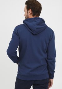 Sötétkék kapucnis pulóver, húzózsinórral, bordás mandzsettákkal és aljjal. A bal ujjaon logó folt található, sima, texturált anyagból készült.