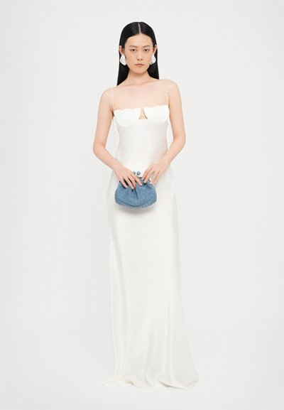 Rochie albă din satin cu decolteu adânc și accente florale. Ține în mână o geantă de umăr texturată, de culoare albastră, cu un design răsucit.