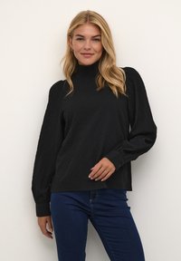 Černý pletený pulovr s vysokým límcem, dlouhými rukávy a zapínáním na knoflíky. Kombinovaný s tmavými džínami, představený na neutrálním pozadí.