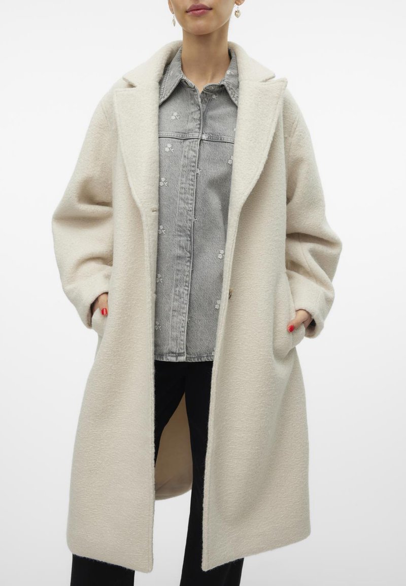 Crèmekleurige, textuurjasje met een ontspannen pasvorm; heeft een brede kraag, zijzakken en een middellange stijl, gelaagd over een grijze shirt.