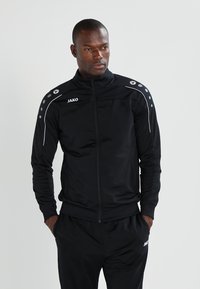Veste de sport noire avec une fermeture éclair à l'avant, présentant des accents blancs et des points aux épaules. Fabriquée dans un tissu lisse avec une coupe ajustée.