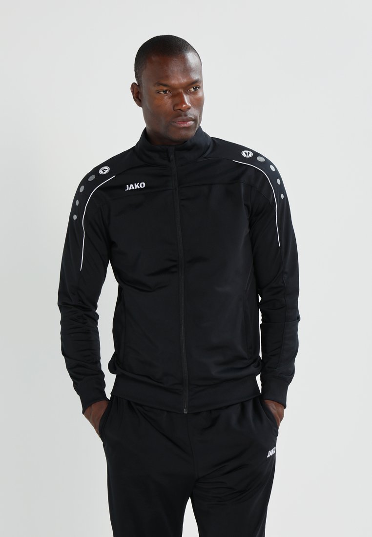 Veste de sport noire avec une fermeture éclair à l'avant, présentant des accents blancs et des points aux épaules. Fabriquée dans un tissu lisse avec une coupe ajustée.