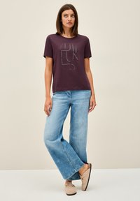 Musta purppura t-paita, jossa on graafinen kuvio, yhdistettynä vaaleansinisiin leveälahkeisiin farkkuihin ja beigen värisiin liukuvataviin kenkiin. Suhtsmooth-kankaan rakenne.