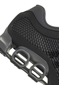 Zapatilla deportiva negra con parte superior de malla, acentos de goma suave y una suela texturizada con elementos de amortiguación visibles y ojales.