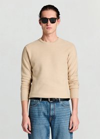 Beiger gerippter Pullover mit langen Ärmeln und rundem Ausschnitt, kombiniert mit blauen Jeans und einem schwarzen Gürtel, ergänzt durch eine schwarze Sonnenbrille.