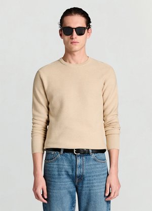Maglione beige a coste con maniche lunghe e scollatura rotonda, abbinato a jeans in denim blu e cintura nera, completato da occhiali da sole neri.