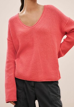 Femme portant un pull texturé rose corail à manches longues avec un col rond et un pantalon noir ample, main posée sur la hanche.